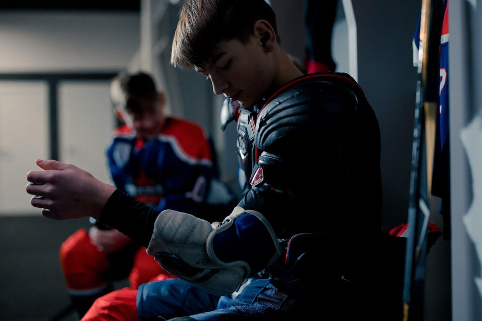 A hockey player gearing up for the ice by stapping on his elbow pads and shoulder pads