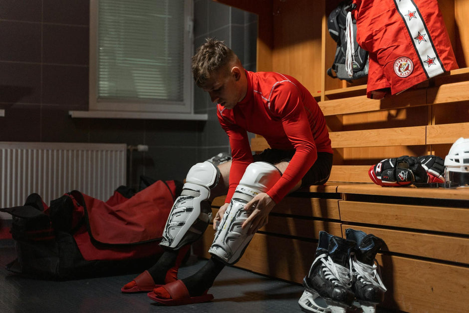 A hockey player strapping on shin guards and other hockey accessories with hockey skates, hockey gloves and a hockey helmet in the background